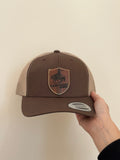 Brown and beige cap with a logo held by a hand against a beige background